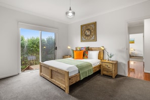 A bedroom with a wooden bed, a sliding glass door to a garden, a bedside table with a lamp, and a wall-mounted artwork. - Tumby on Rye (Rye)