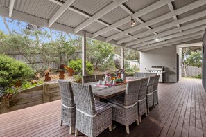 An outdoor patio with a dining table and chairs, surrounded by greenery and a barbecue grill.