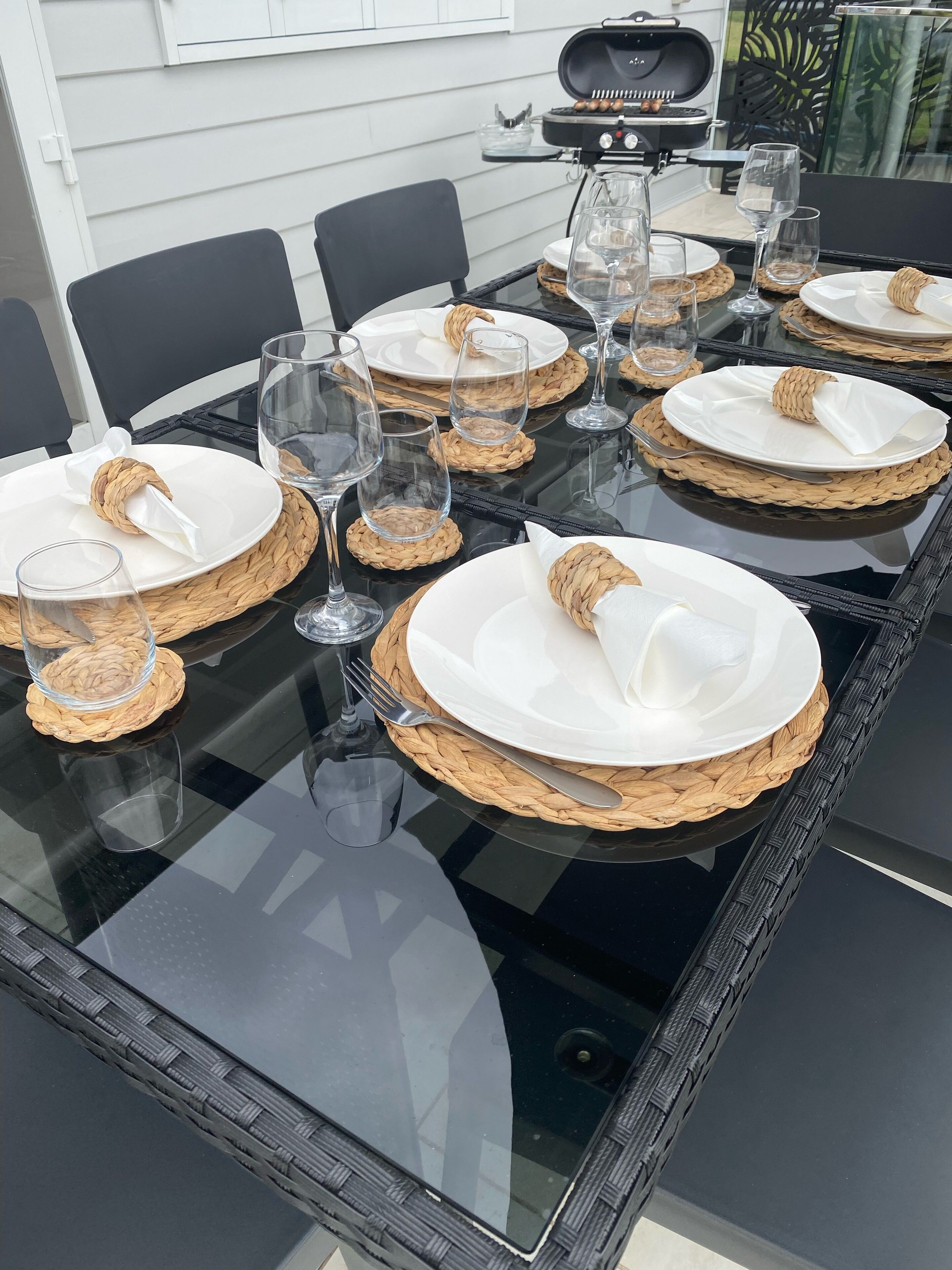 Une table à manger dressée avec de l’argenterie blanche, des sets de table en fibres tressées et de la verrerie transparente.