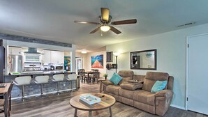 A spacious living room with a brown leather sofa, a wooden coffee table, and a ceiling fan. A kitchen with white cabinets and a bar area is visible in the background. - Big-Screen Ducks Nights, Autzen Stadium 4 Mi Away (Springfield)