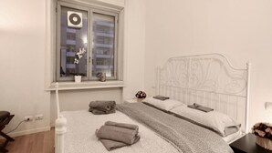 Quarto com cama, janela com vista para edifícios, mesa de cabeceira com um vaso de flores e ar-condicionado fixado na parede.