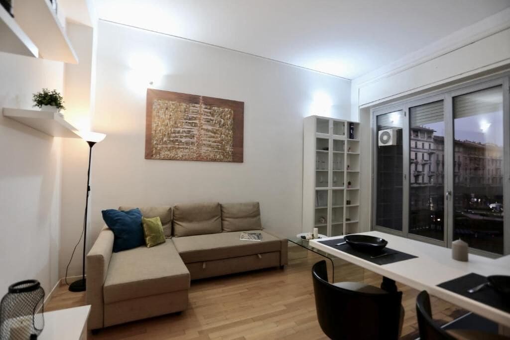 A modern living room with a sofa, a dining table, and a large window with a city view.