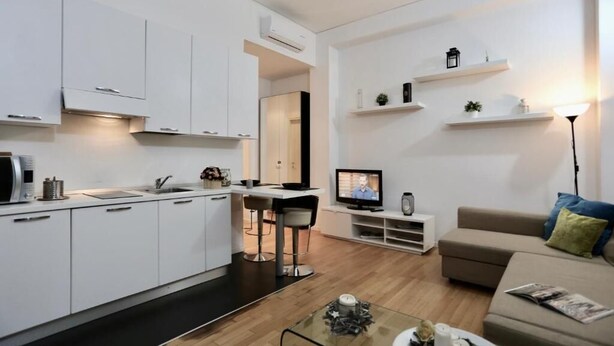 Uma cozinha moderna com armários brancos, uma área de jantar com mesa e cadeiras, uma televisão e um sofá em uma sala de estar.