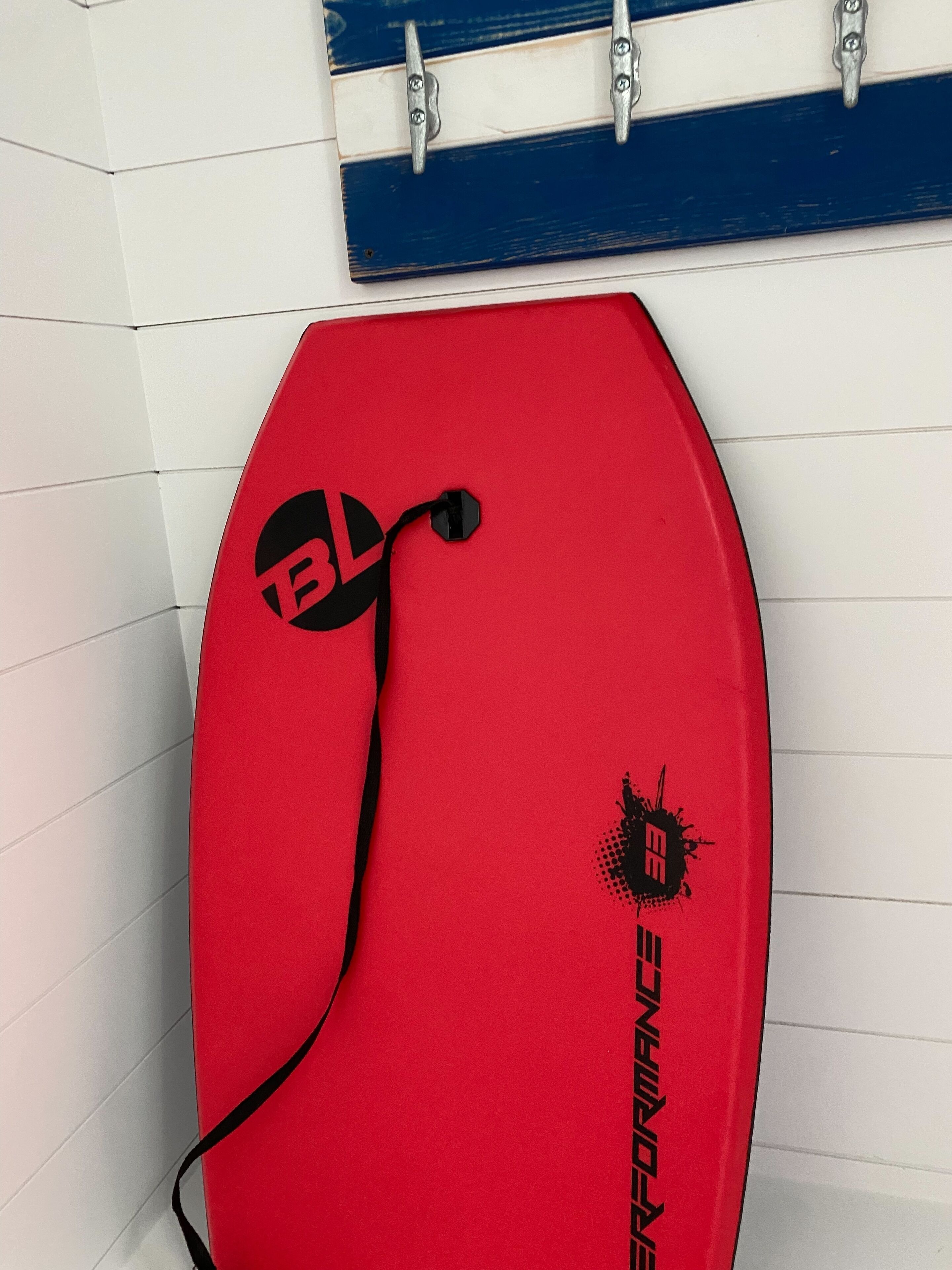 A red surfboard with a logo and a black strap, leaning against a tiled wall.