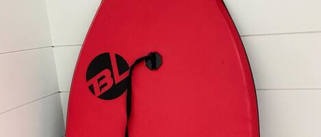 A red surfboard with a logo and a black strap, leaning against a tiled wall.
