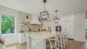 A modern kitchen with white cabinetry, a large island with bar stools, pendant lighting, and a view of greenery outside. - Private Home with Custom Pool and Spa! (Bradenton Beach)