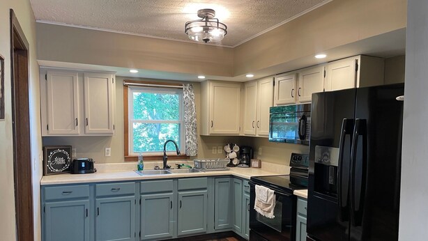 A kitchen with light blue cabinets, a black refrigerator, and a window with a view of trees. - The Everett House (O'Neill)