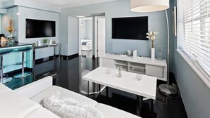 A modern hotel room with a dining area, a large flat-screen TV, a white sofa, and a large window with blinds.