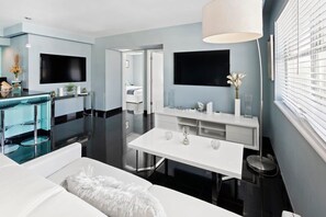 A modern hotel room with a dining area, a large flat-screen TV, a white sofa, and a large window with blinds.