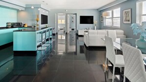 A modern kitchen with a central island, a dining area with white chairs, and a living space with a sofa and a TV.