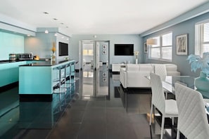 A modern kitchen with a central island, a dining area with white chairs, and a living space with a sofa and a TV.