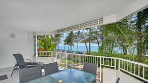 A balcony with a glass table and chairs, overlooking a beach with palm trees and a view of the ocean. - Costa Royale 3 Bedroom Beachfront Apartment (Trinity Beach)