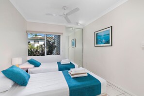 A hotel room with two beds, a ceiling fan, a window with a view, and a framed picture on the wall. - Costa Unit 1 | 3 Bedroom Beachfront Apartment (Trinity Beach)