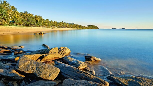 A beach with clear water, rocks in the foreground, and a forested area in the background. - Costa Royale 3 Bedroom Beachfront Apartment (Trinity Beach)