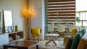 Un salon moderne avec un canapé, une table basse et un coin repas avec des chaises. On y trouve une grande fenêtre avec des stores, une lampe sur pied et un vase décoratif posé sur une étagère.