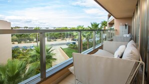 Un balcon donnant sur une route et sur un espace verdoyant, un canapé blanc avec des coussins et une rambarde en verre.