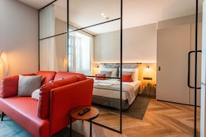 A modern hotel room with a red sofa, a bed with white bedding, a small round table with a remote control, and a large mirror reflecting the room.