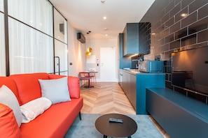 A modern living room with a red sofa, a small round table, and a kitchen area with a blue countertop and a microwave.