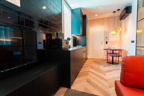 Une cuisine moderne avec des meubles de rangement noirs, un parquet en bois disposé en chevrons, un coin repas avec une chaise rouge et des luminaires suspendus.