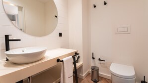 Un baño moderno con un lavamanos blanco, un espejo redondo, un inodoro y un cubo de basura.