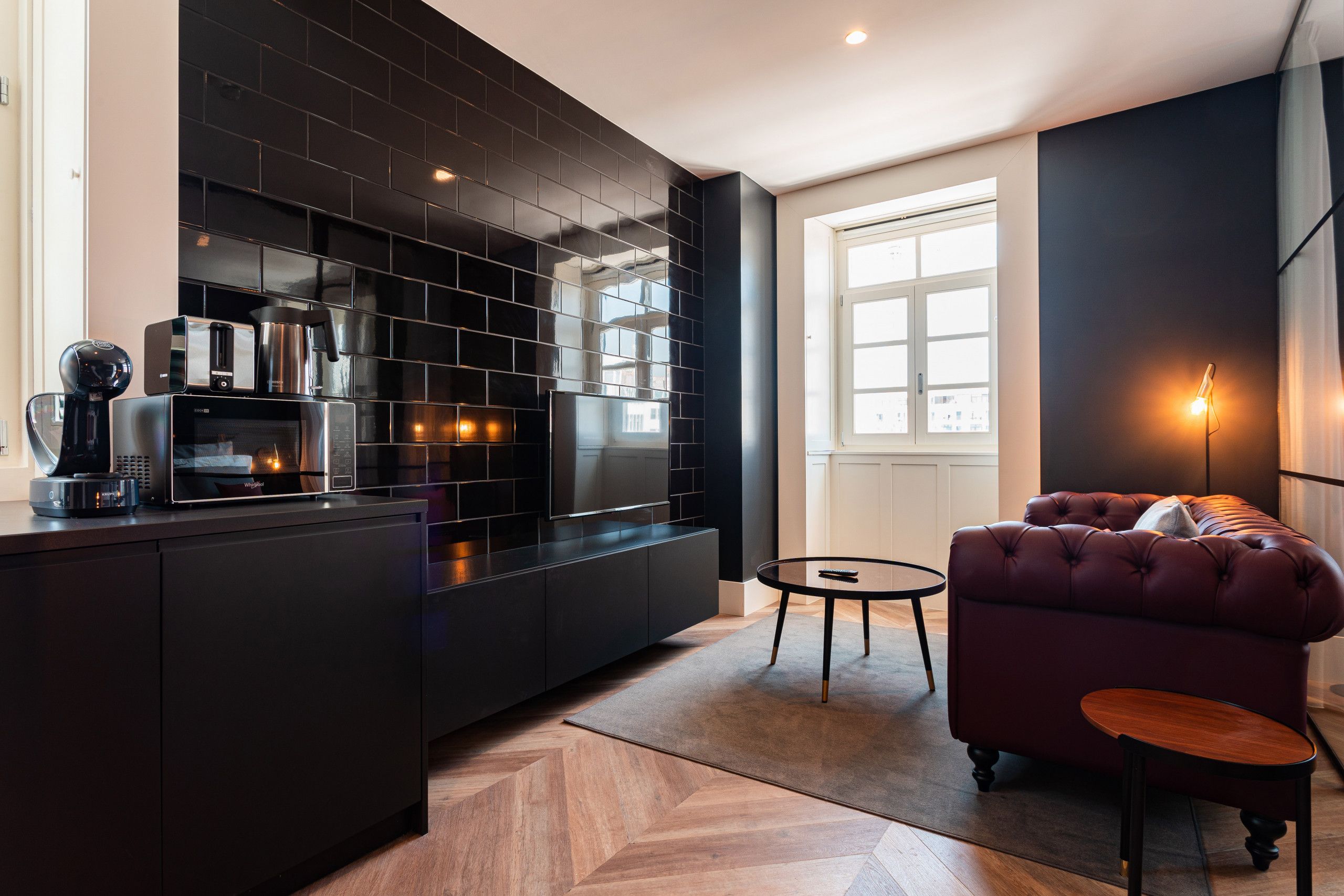 A modern kitchen with black cabinetry, a built-in oven, and a coffee machine. There is a round table and a tufted sofa with a small side table.