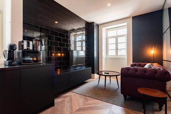 A modern kitchen with black cabinetry, a built-in oven, and a coffee machine. There is a round table and a tufted sofa with a small side table.