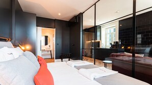 Ein modernes Hotelzimmer mit einem großen Bett, einer Sitzecke mit einer Couch und einer Glaswand, die Blick auf die Küche und den Speisebereich bietet.
