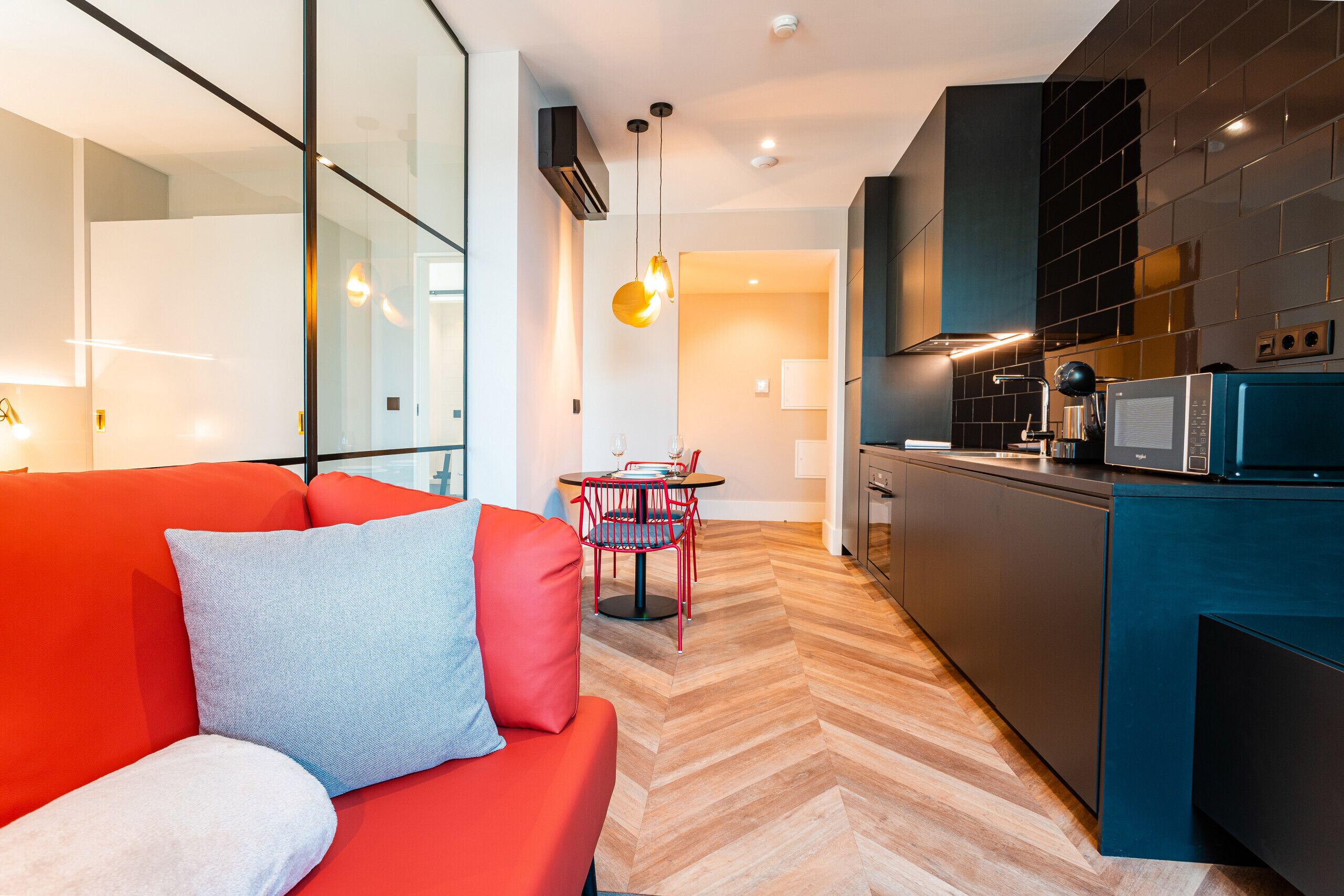 A modern living room with a red sofa, a small dining table with a red chair, and a kitchen area with dark cabinetry and a microwave.