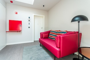 A red sofa with a colorful pillow, a black floor lamp, and a red fire alarm on the wall.
