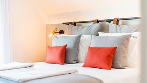 Una cama bien hecha con ropa de cama blanca y gris, un cabecero con una lámpara y una ventana con cortinas blancas.