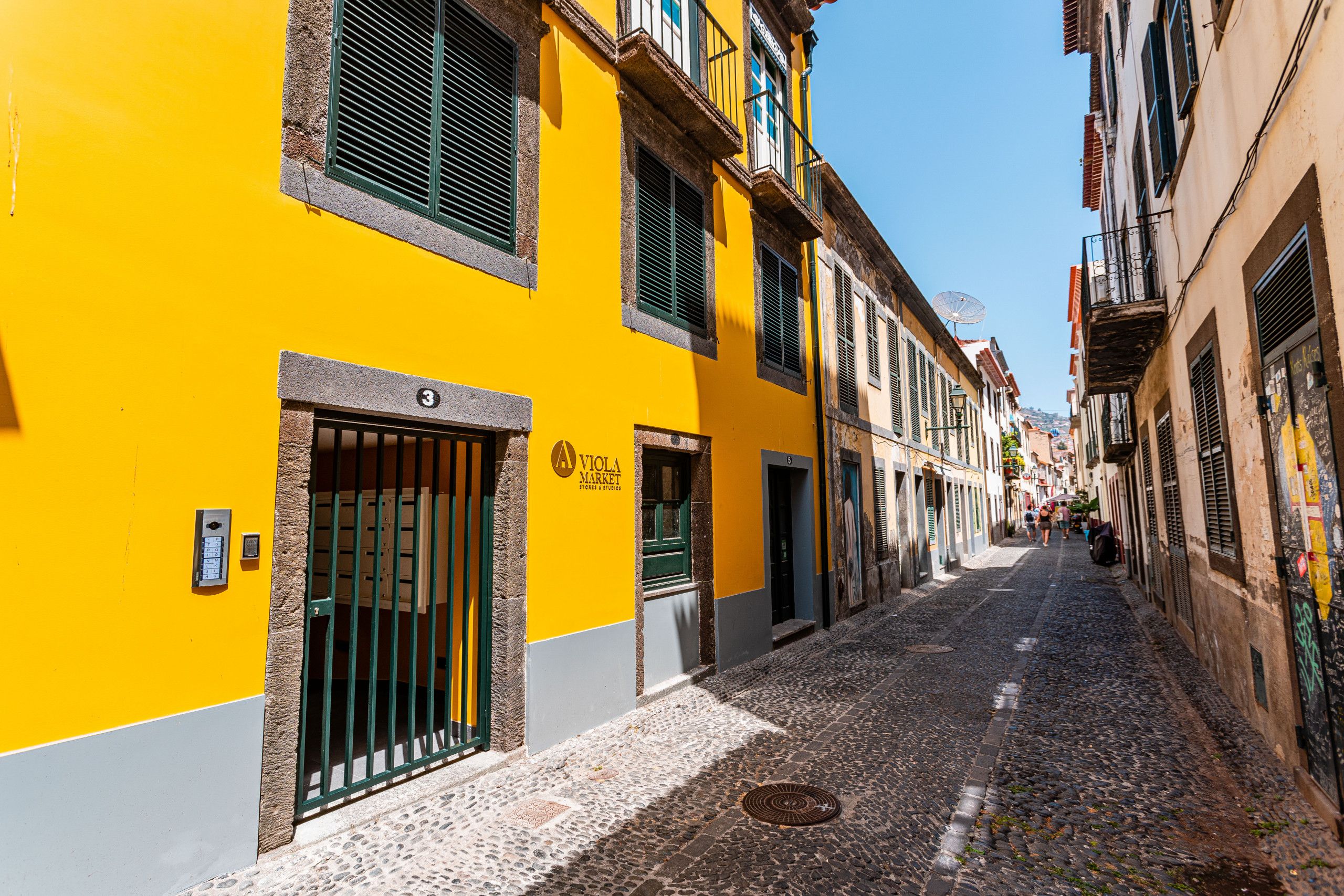A narrow cobblestone street lined with yellow buildings, each with a distinct architectural style and visible doorways.