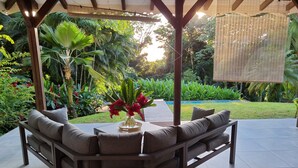 A covered outdoor seating area with a sofa and a table, surrounded by lush greenery and a small pool. - Villa with exceptional view of tropical garden! (Rivière-Salée)