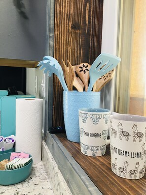 Un comptoir de cuisine sur lequel se trouve un récipient bleu contenant des ustensiles en bois, un rouleau d’essuie-tout et deux tasses décorées de motifs représentant un chien et un lama.