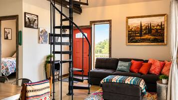 A modern living room with a black spiral staircase, a red door, a black sofa with colorful cushions, and a large landscape painting on the wall.