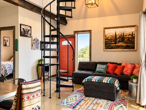 Un salon moderne avec un escalier en colimaçon noir, une porte rouge, un canapé noir agrémenté de coussins colorés et un grand tableau représentant un paysage accroché au mur.