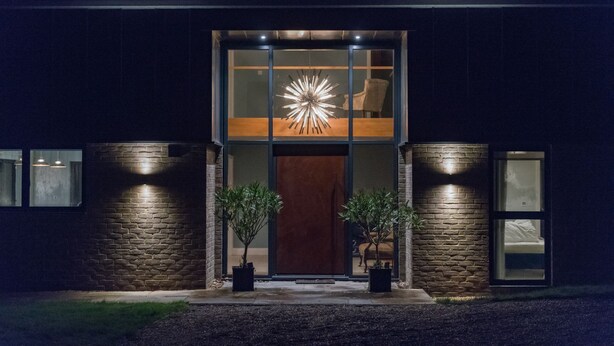 Un edificio moderno con una amplia entrada de vidrio, dos plantas en macetas y un aplique de luz único sobre la puerta.
