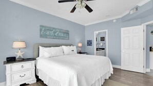 A bedroom with a bed, nightstands, a ceiling fan, a wall with a 'beach dreams' sign, and a door leading to another room. - San Carlos 1407 by Liquid Life (Gulf Shores)