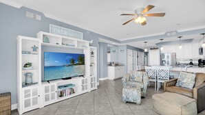A spacious living room with a large flat-screen TV, white built-in shelving, a ceiling fan, and a kitchen area with white cabinets and a dining table. - San Carlos 1407 by Liquid Life (Gulf Shores)