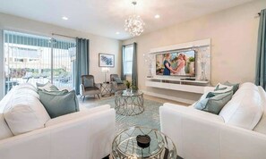 A modern living room with a flat-screen TV, white sofas, a glass coffee table, and a chandelier. - Sleeps 30 | 11BR Pool Home w/ Theater & Game Room (Kissimmee)
