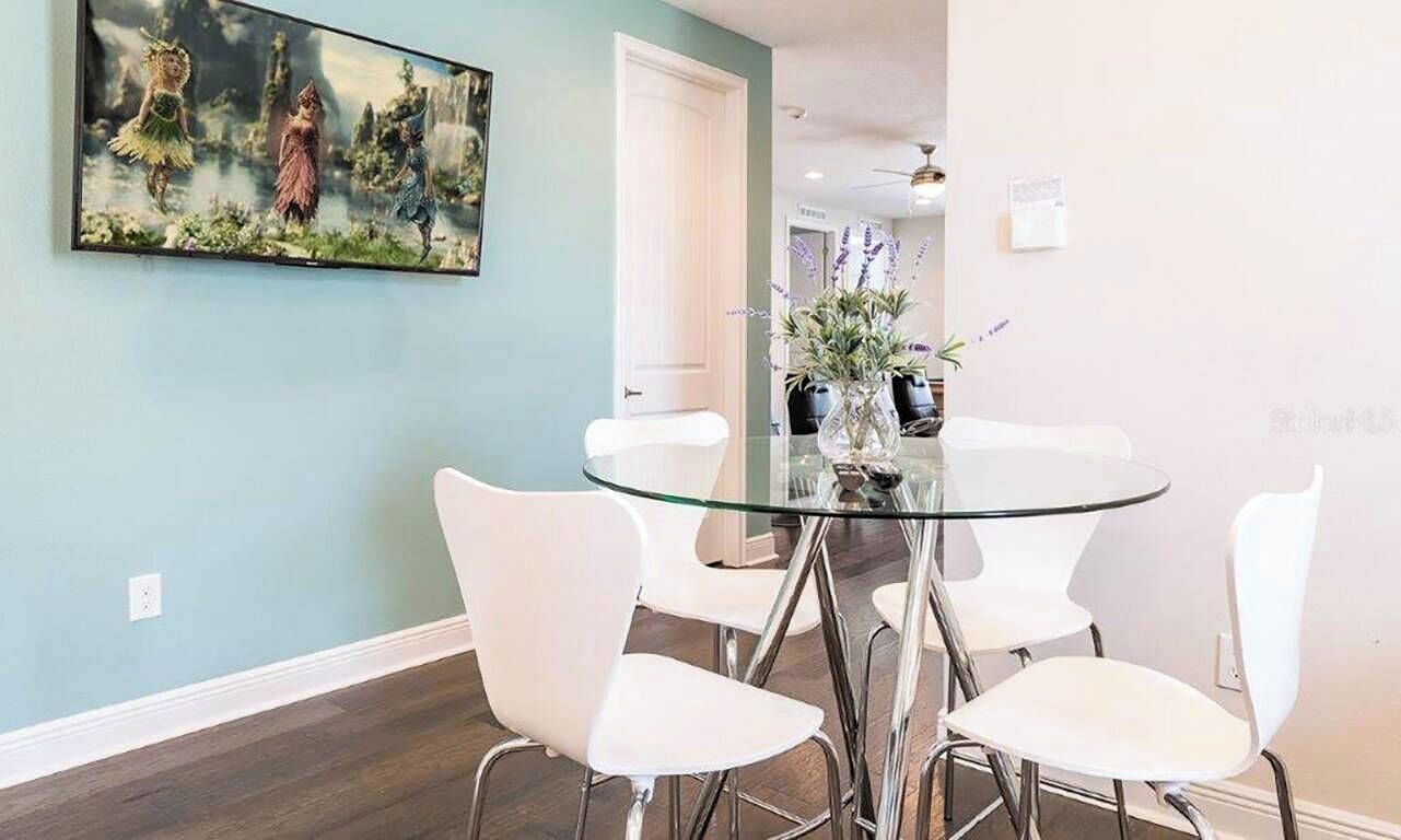A dining area with a glass table surrounded by white chairs, a painting on the wall, and a vase with flowers on the table.
