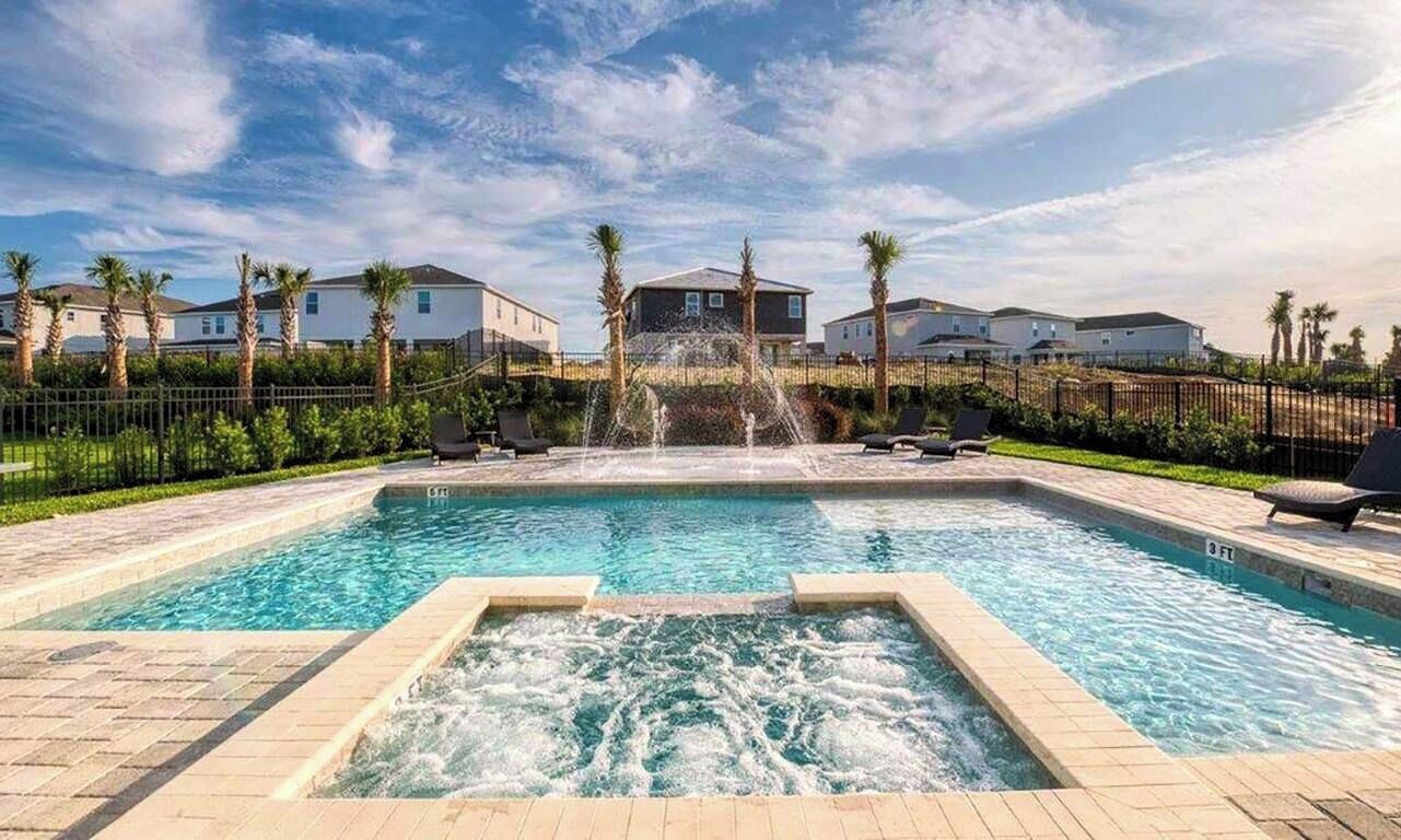 A swimming pool with a fountain feature, surrounded by palm trees and a fence, with houses in the background.