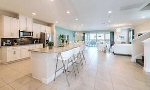 A modern kitchen with white cabinetry, a central island with bar stools, and a view to a living area with a staircase. - Sleeps 30 | 11BR Pool Home w/ Theater & Game Room (Kissimmee)