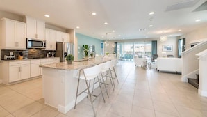 A modern kitchen with white cabinets, a granite countertop, and stainless steel appliances. There are bar stools and a dining area with a white table and chairs. - Encore Resort 11 Bedroom Home with Pool (2117) (Kissimmee)