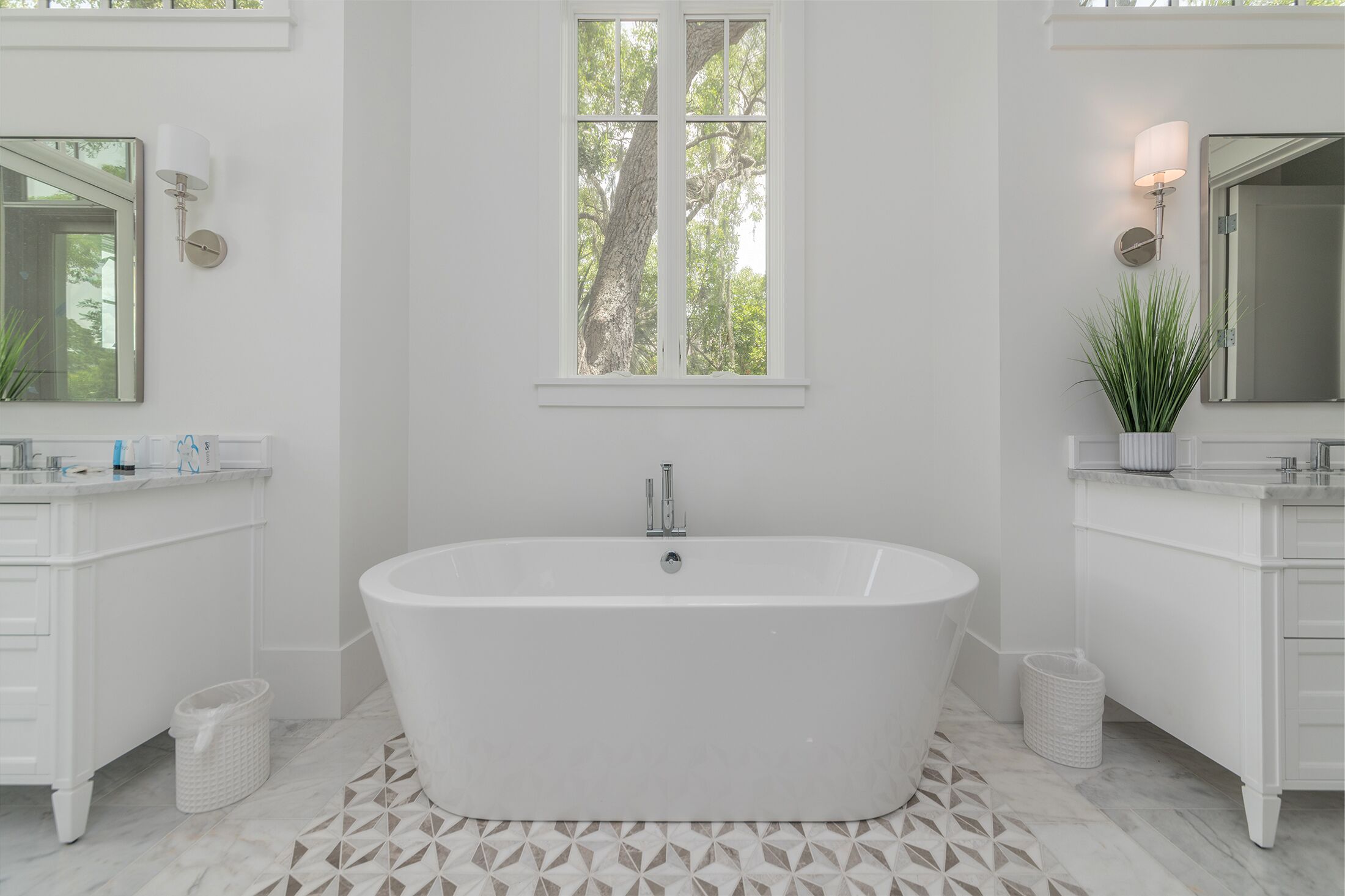 Un baño moderno con una bañera independiente, dos lavamanos y un amplio ventanal con vistas a árboles, además de una planta en maceta.