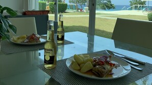 A dining table with a plate of food, two bottles of Corona Extra beer, and a glass. The table is near a large window with a view of a beach and umbrellas.