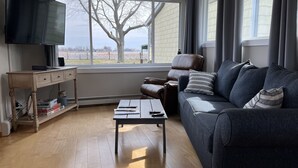 A living room with a leather sofa, a coffee table, a TV on a stand, and a window with a view of trees and a building. -  Cabana Bay Fish Camp, Lake St. Clair- Minutes to Anchor Bay (Clay Township)