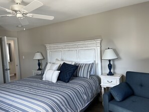Quarto com cama, duas mesas de cabeceira, uma cadeira, ventoinha no teto e uma porta.