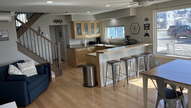 A modern kitchen with a central island, a dining area with bar stools, a staircase leading to another level, and a view of the outside through a large window. -  Cabana Bay Fish Camp, Lake St. Clair- Minutes to Anchor Bay (Clay Township)