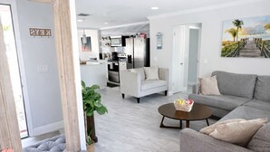 A modern living room with a grey sofa, a grey tufted ottoman, and a coffee table. There is a kitchen in the background with a white fridge and a dining area with a white table and chairs. - Paradise Palms Beach house with HEATED POOL🏖🌅🐬 (Largo)