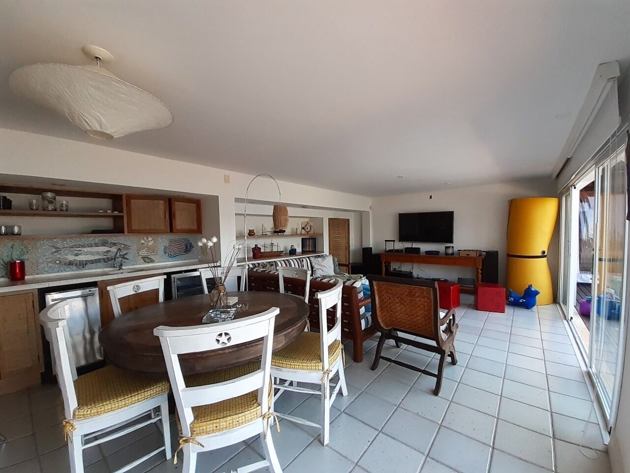 A spacious living area with a dining table surrounded by chairs, a kitchen area with cabinets and a countertop, and a large window providing natural light.